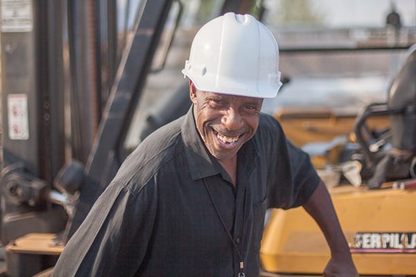 A participant in Avivo's programs wears a hard hat and stands near a forklift at a construction site.